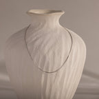 The Dainty Herringbone Necklace in Sterling Silver drapes elegantly over a textured, off-white ceramic vase for a refined layered look.
