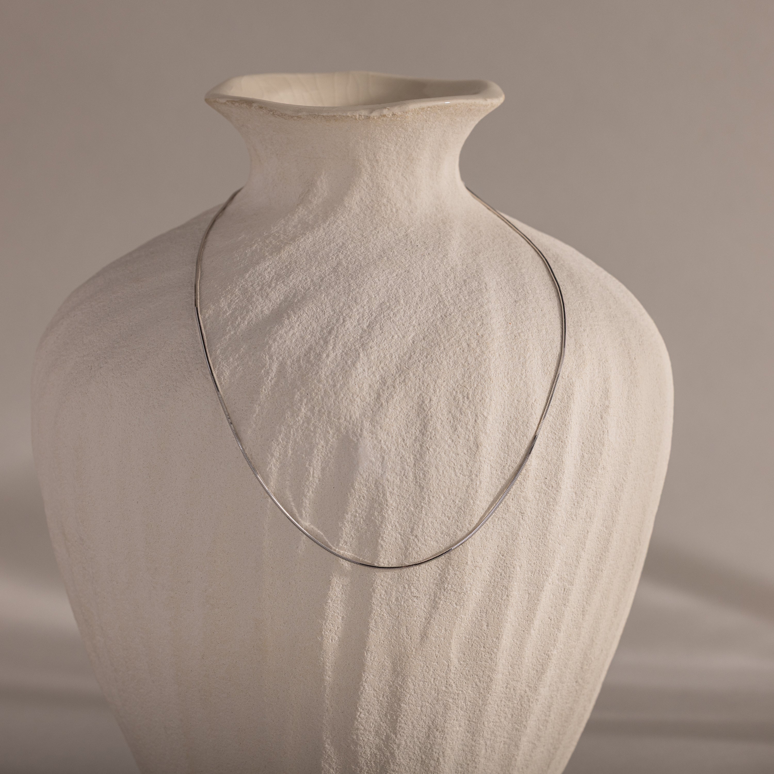 The Dainty Herringbone Necklace in Sterling Silver drapes elegantly over a textured, off-white ceramic vase for a refined layered look.
