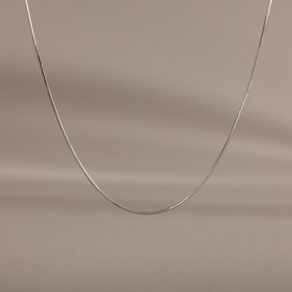 The Dainty Herringbone Necklace in Sterling Silver is displayed on a neutral beige background, ideal for adding a subtle shine to your layered look.