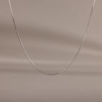 The Dainty Herringbone Necklace in Sterling Silver is displayed on a neutral beige background, ideal for adding a subtle shine to your layered look.