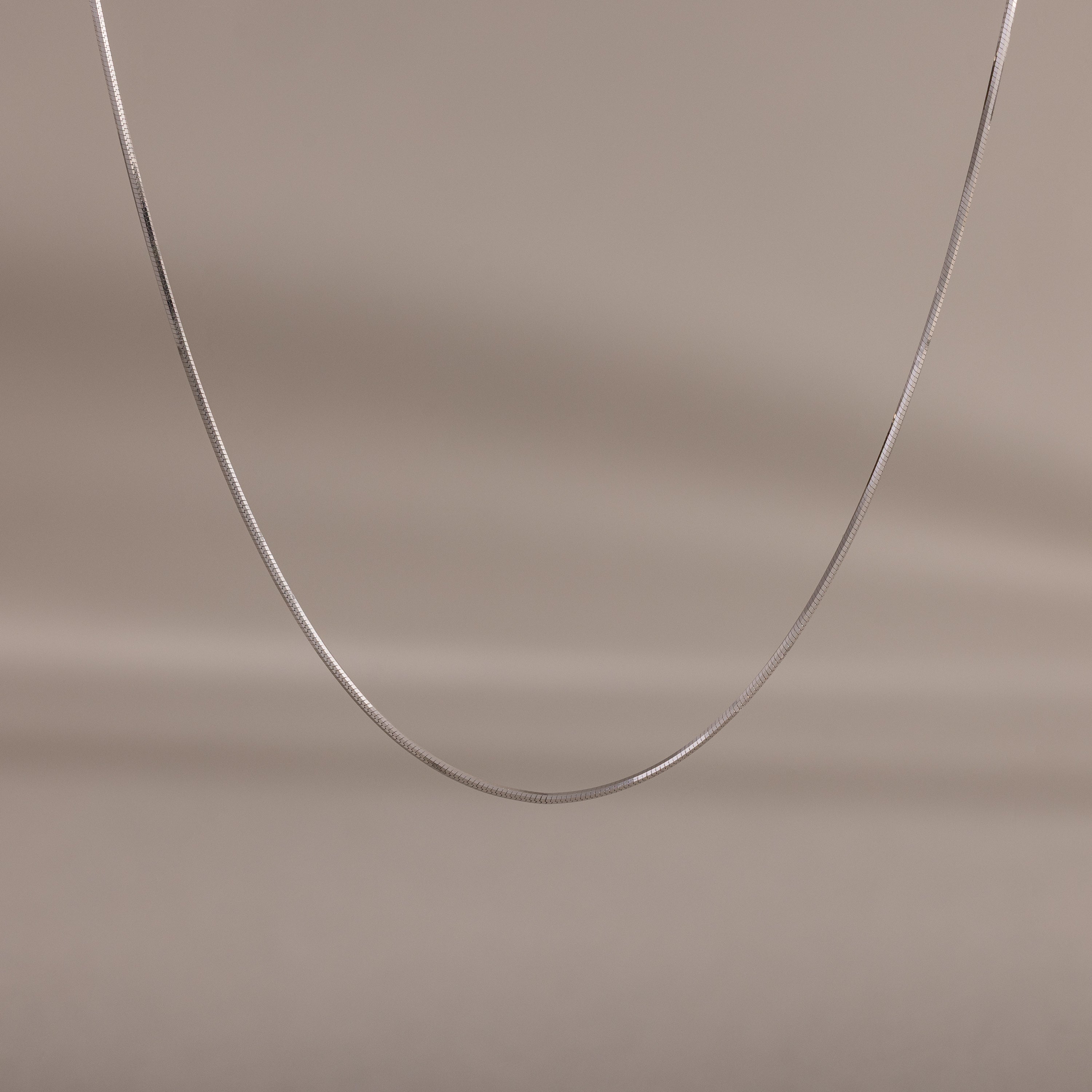 The Dainty Herringbone Necklace in Sterling Silver is displayed on a neutral beige background, ideal for adding a subtle shine to your layered look.
