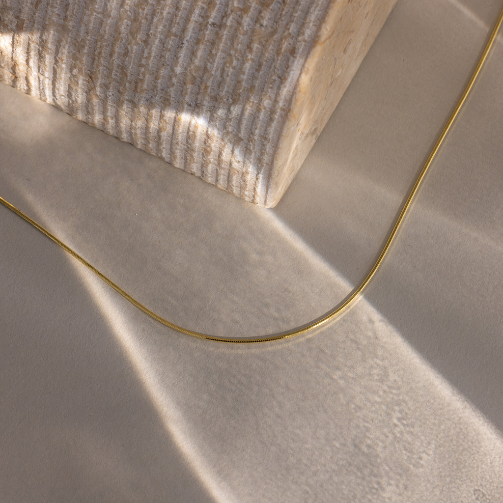 The Dainty Herringbone Necklace in 18K Gold rests on a textured beige surface next to a ridged stone block, illuminated by soft light.