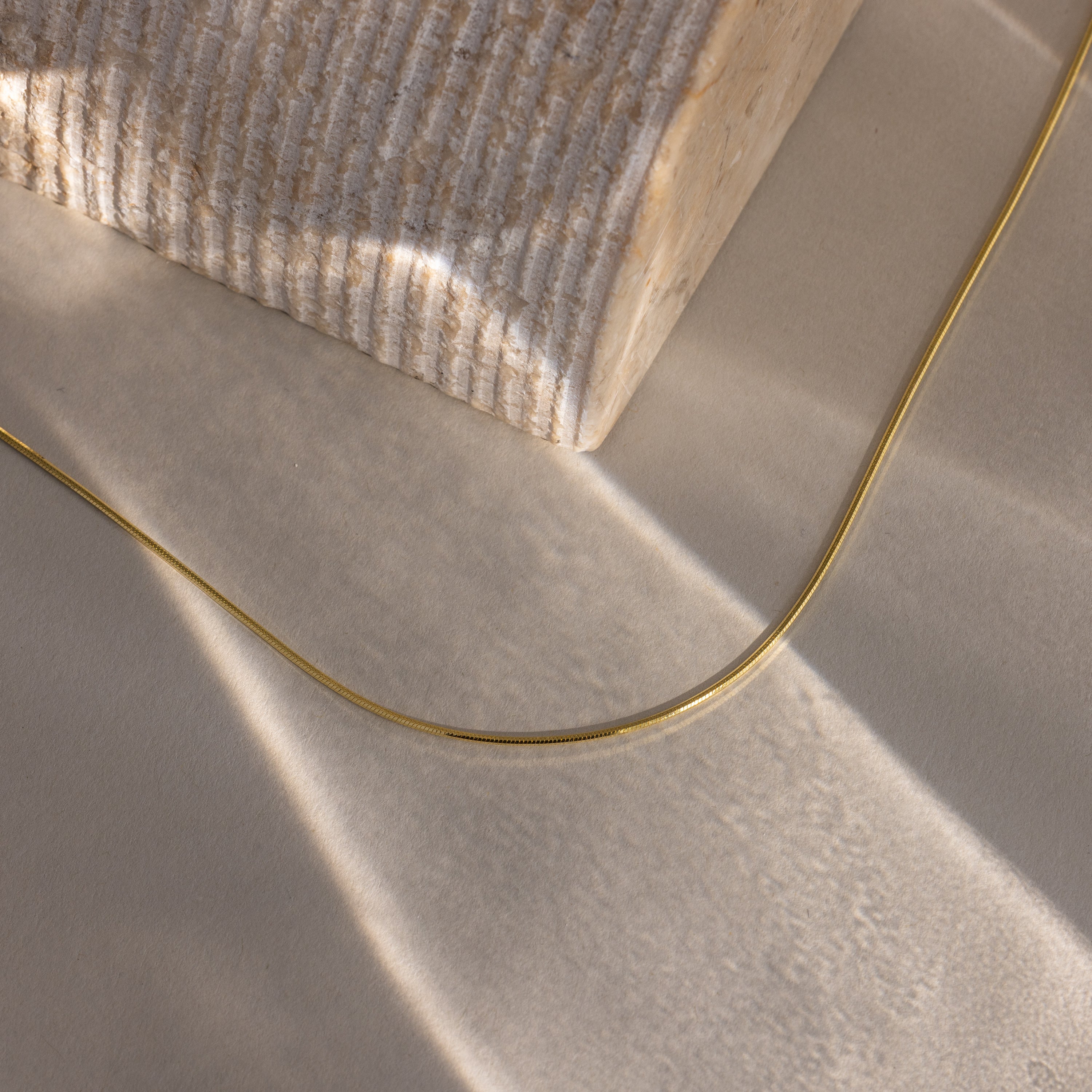 The Dainty Herringbone Necklace in 18K Gold rests on a textured beige surface next to a ridged stone block, illuminated by soft light.