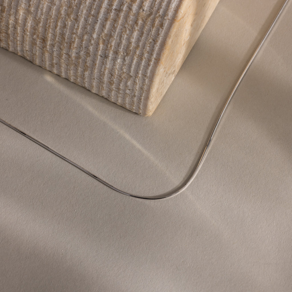The Dainty Herringbone Necklace is displayed resting on a beige surface beside a textured block.