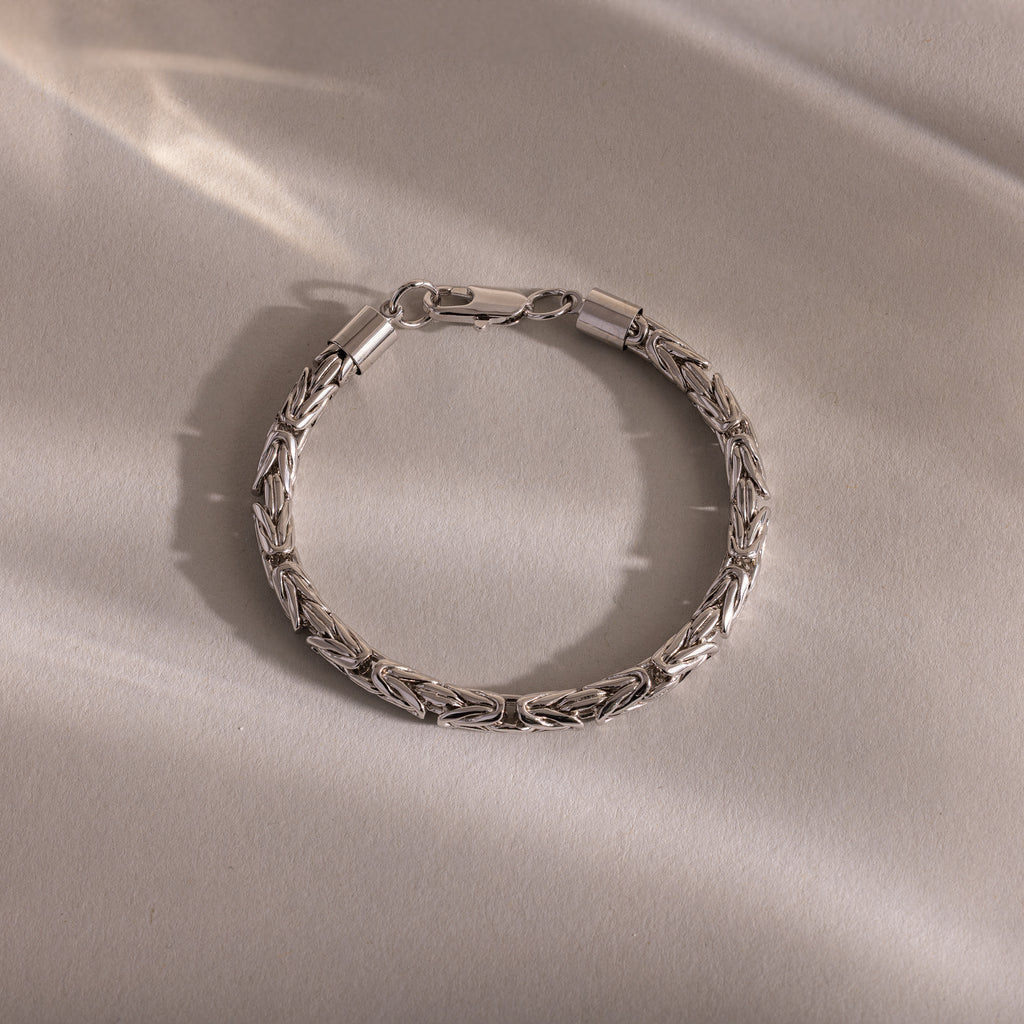 The Byzantine Link Bracelet in White Gold features an intricate interwoven pattern and rests on a light beige surface, its detailed links casting soft shadows—a striking statement jewelry piece.