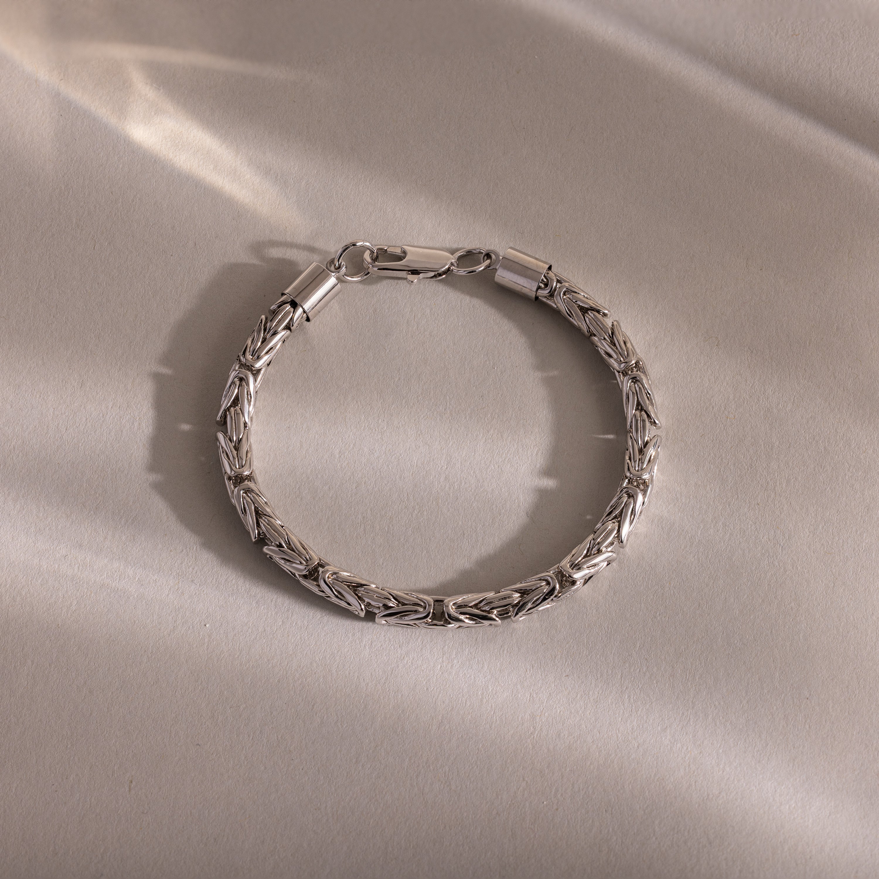 The Byzantine Link Bracelet in White Gold features an intricate interwoven pattern and rests on a light beige surface, its detailed links casting soft shadows—a striking statement jewelry piece.