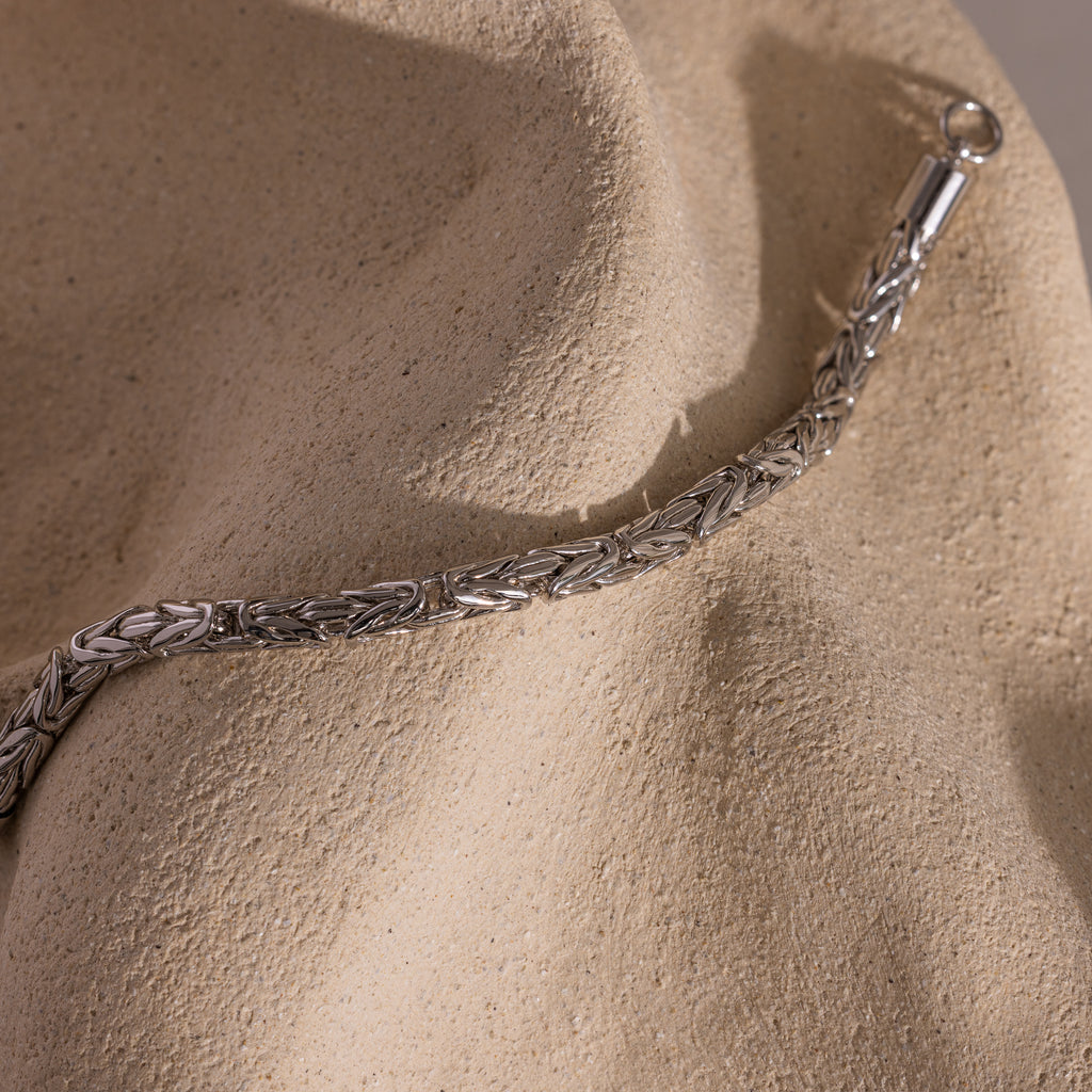 The Byzantine Link Bracelet in White Gold features an interwoven pattern and is displayed on a textured beige stone surface—ideal as statement jewelry.