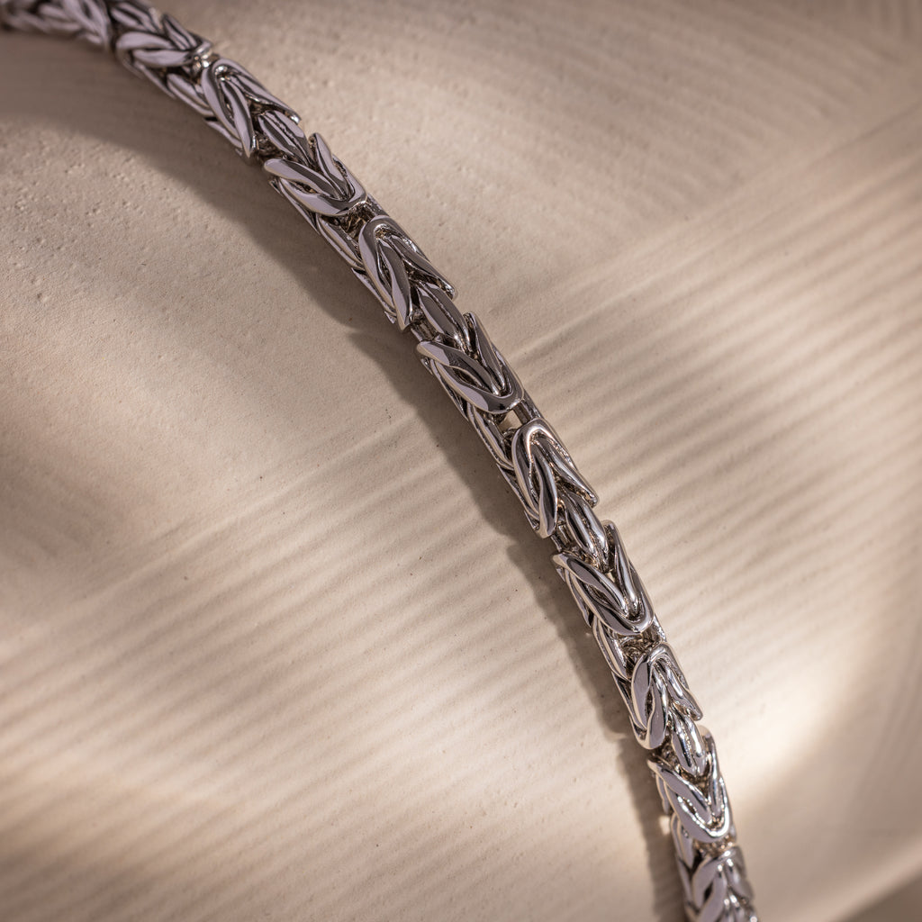 The Byzantine Link Bracelet, featuring a silver interwoven leaf pattern, rests on a textured beige surface, casting soft shadows and offering a subtle statement look.