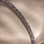 The Byzantine Link Bracelet, featuring a silver interwoven leaf pattern, rests on a textured beige surface, casting soft shadows and offering a subtle statement look.