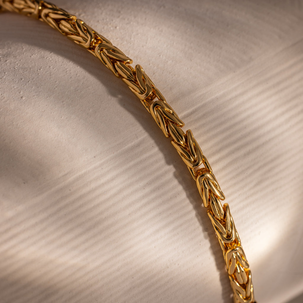 Close-up of the Byzantine Link Bracelet in 18K Gold, featuring an interwoven design and bold statement style, displayed on a beige, textured surface.
