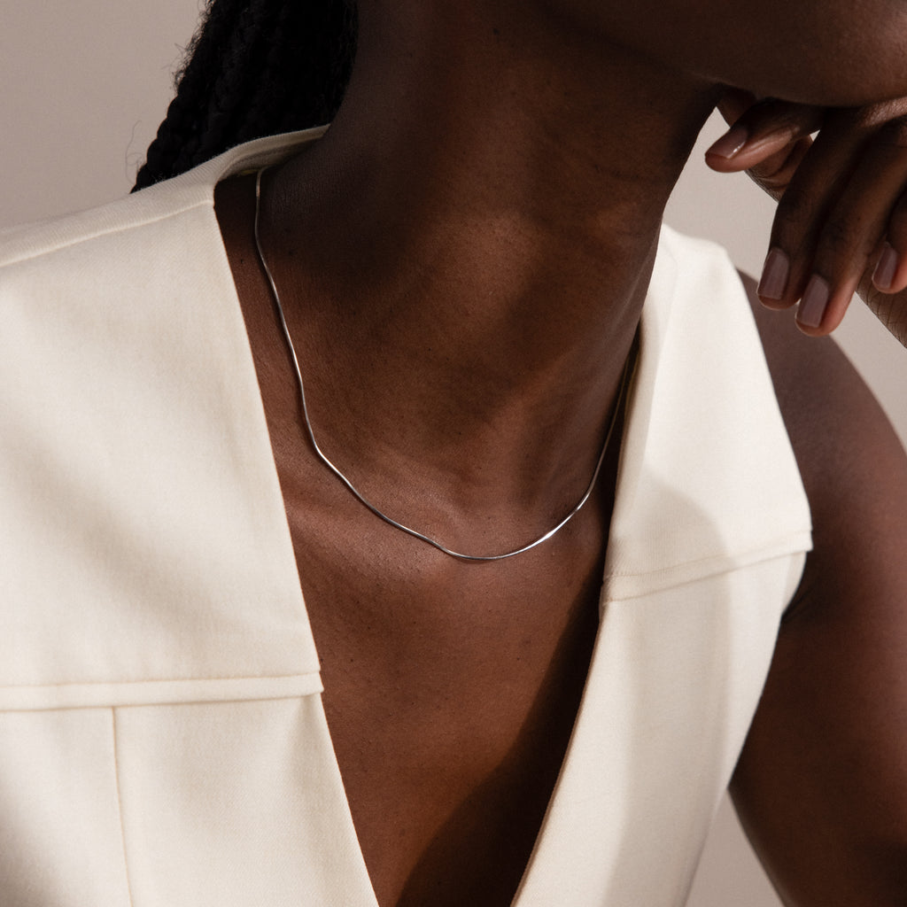 A woman wears a minimalist Dainty Herringbone Necklace in Sterling Silver with an off-white sleeveless top, resting her chin on her hand for a chic, layered style.
