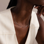 A woman wears a minimalist Dainty Herringbone Necklace in Sterling Silver with an off-white sleeveless top, resting her chin on her hand for a chic, layered style.