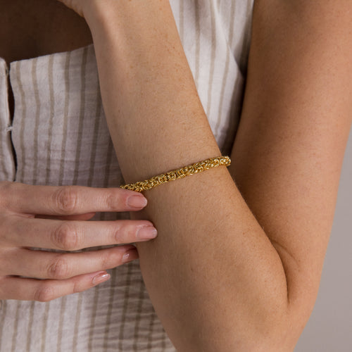 A woman wearing the Bold Byzantine Bracelet in 18K Gold gently touches her arm, dressed in a sleeveless striped top.
