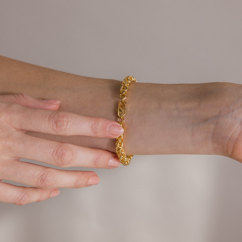 A hand wearing and adjusting the Bold Byzantine Bracelet in 18K Gold with woven links against a neutral background, embodying quiet luxury.