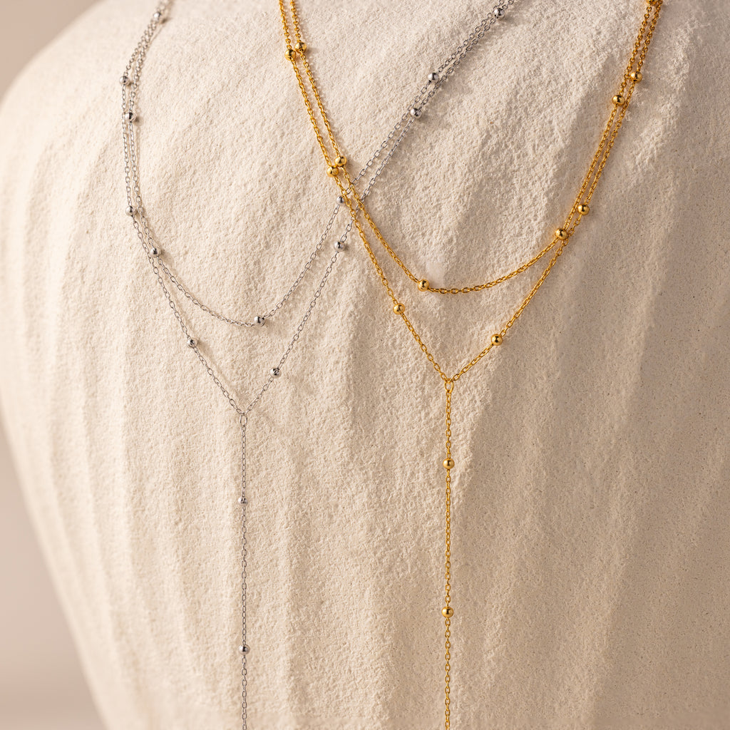 The Duo Satellite Lariat Necklace in Silver, with small beads, is shown layered as minimal jewelry on a textured white fabric background.