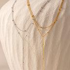 The Duo Satellite Lariat Necklace in Silver, with small beads, is shown layered as minimal jewelry on a textured white fabric background.
