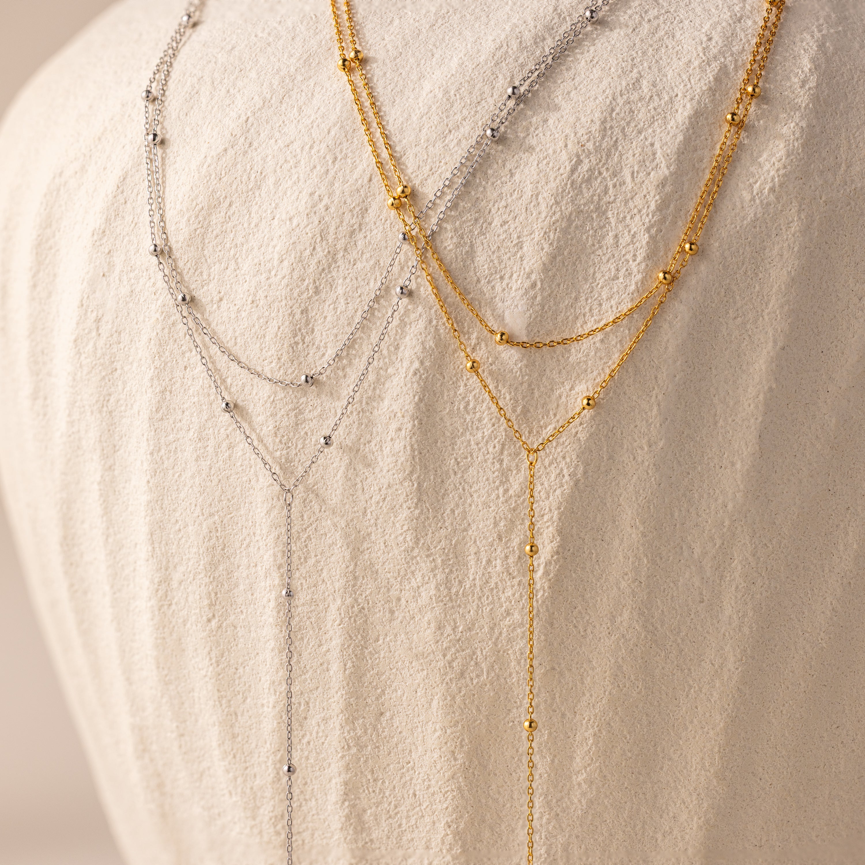 The Duo Satellite Lariat Necklace in Silver, with small beads, is shown layered as minimal jewelry on a textured white fabric background.