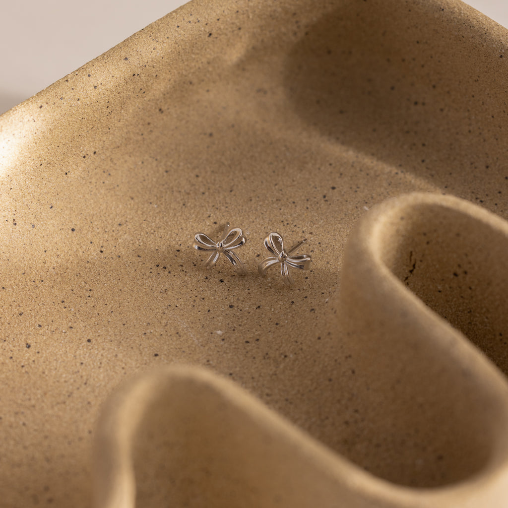 Dainty silver bow stud earrings displayed on a sculpted ceramic tray.