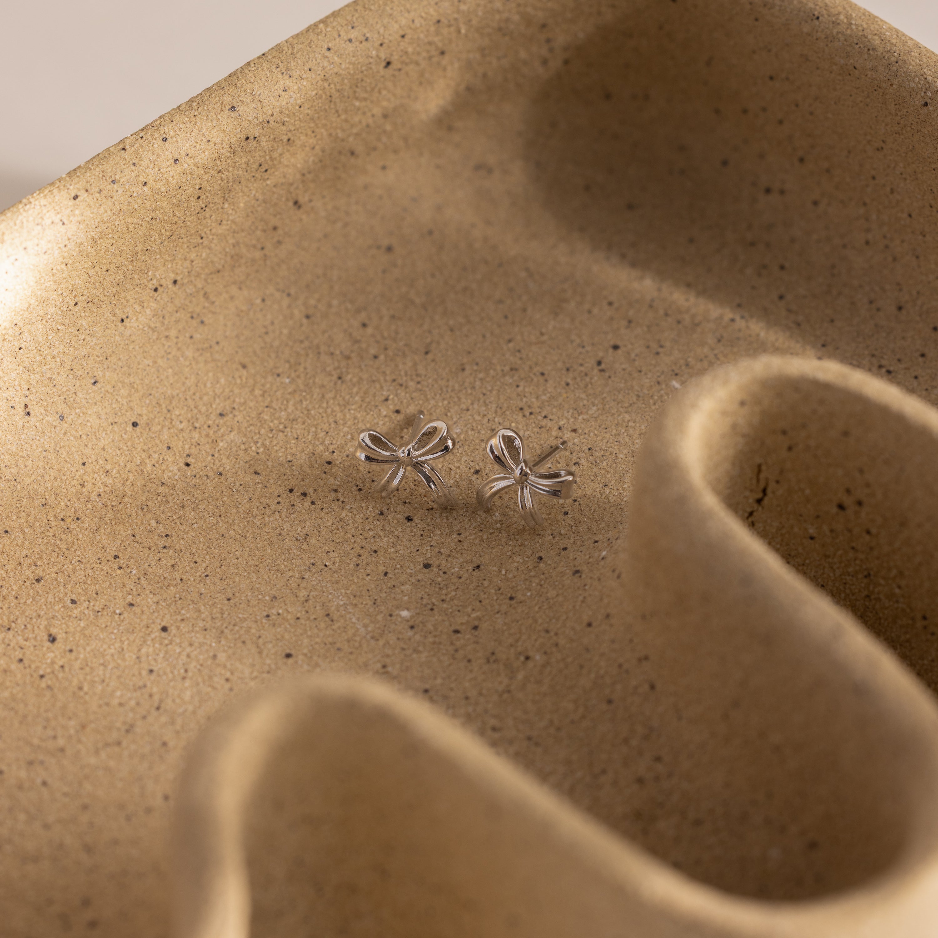 Dainty silver bow stud earrings displayed on a sculpted ceramic tray.