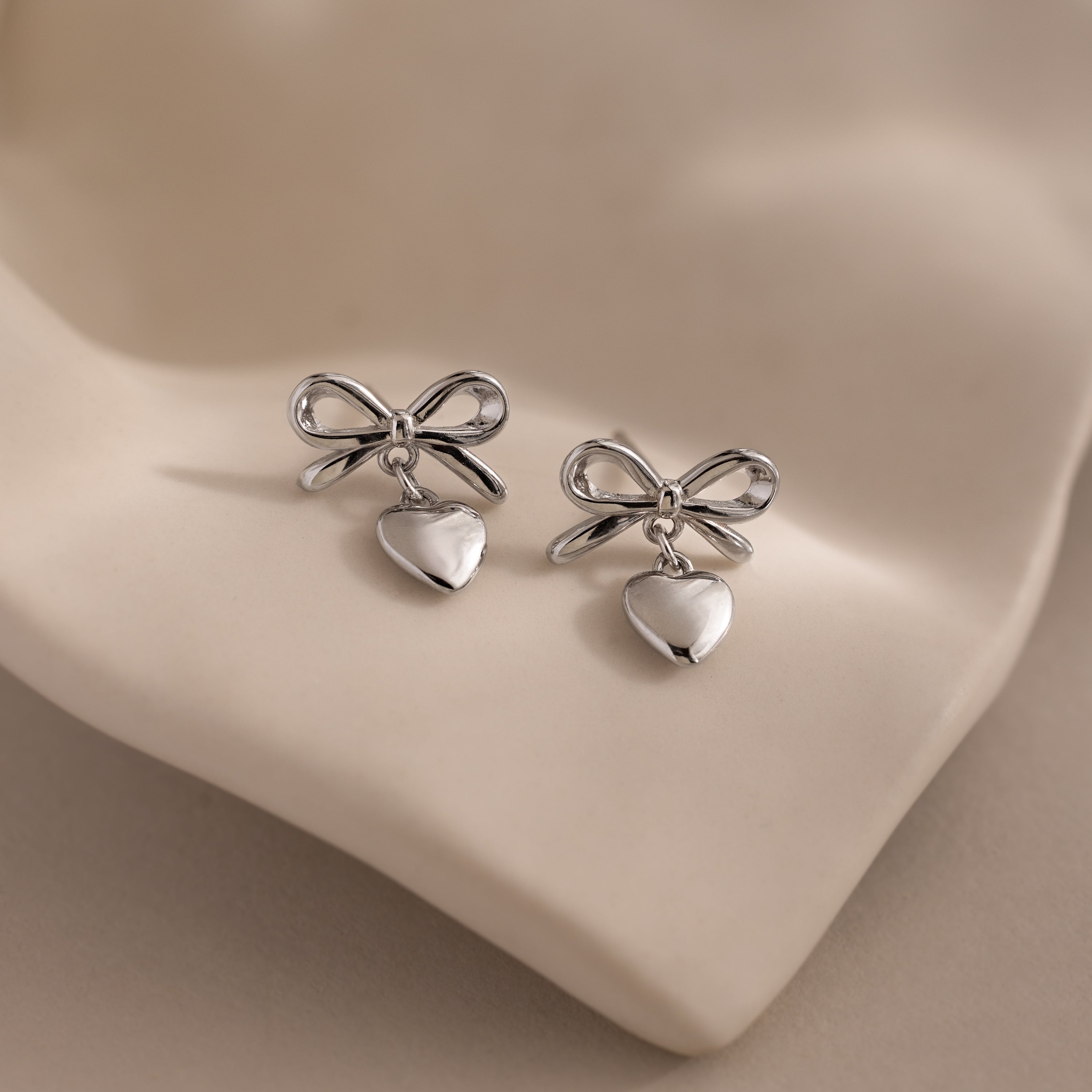 Silver bow earrings with dangling heart charms displayed on a soft, sculpted ceramic dish.