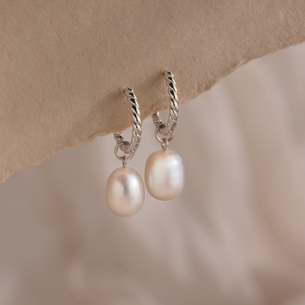 Close-up of elegant silver twisted hoop earrings featuring dangling freshwater pearls, displayed against a soft beige paper background.
