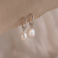 Close-up of elegant silver twisted hoop earrings featuring dangling freshwater pearls, displayed against a soft beige paper background.