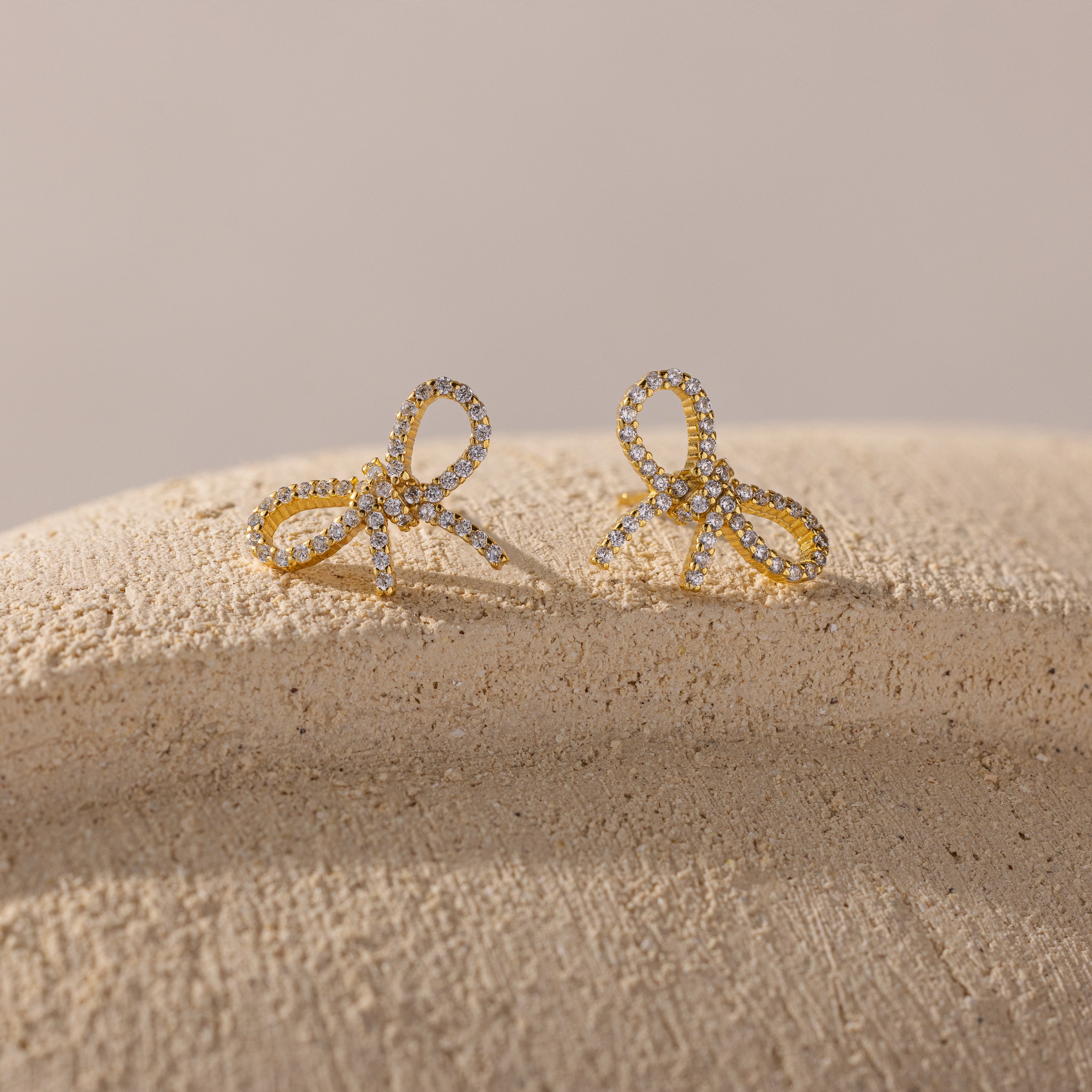 Gold bow-shaped stud earrings encrusted with sparkling cubic zirconia, displayed on a textured neutral surface with soft lighting.