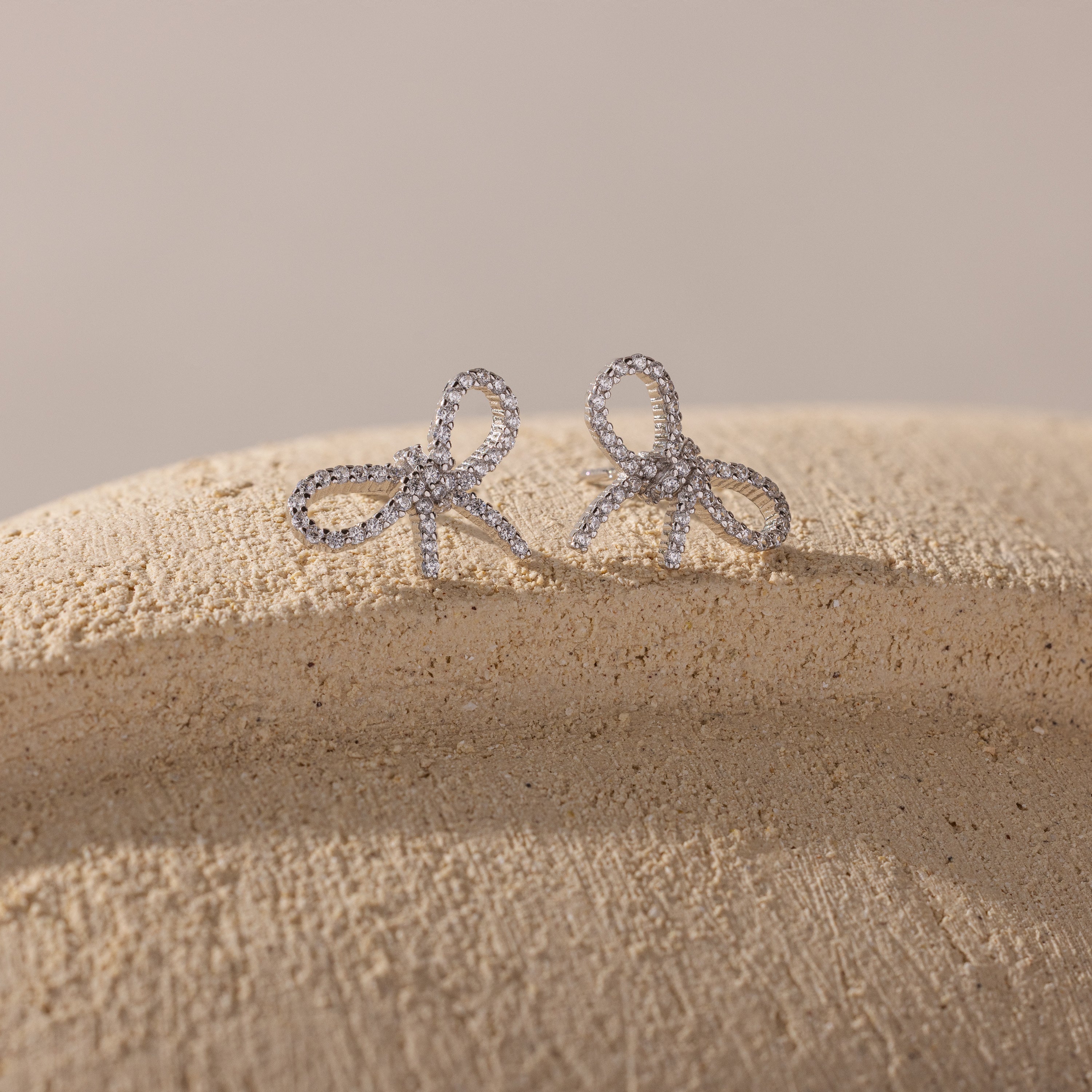 Silver bow-shaped stud earrings adorned with sparkling cubic zirconia, displayed on a textured beige surface with soft natural lighting for a delicate and elegant look.