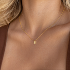 A woman wears a gold Mini Pave Initial Tag Necklace with a small rectangular pendant on her bare neck, highlighting the elegance of personalized jewelry.