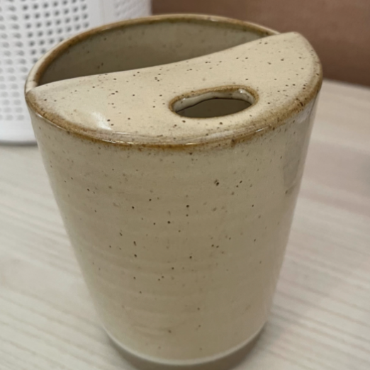 The Free Gift of Choice is a beige ceramic cup with a unique slanted rim and a thumb hole, resting on a light wooden surface—this cute mug brings charm to your daily routine.
