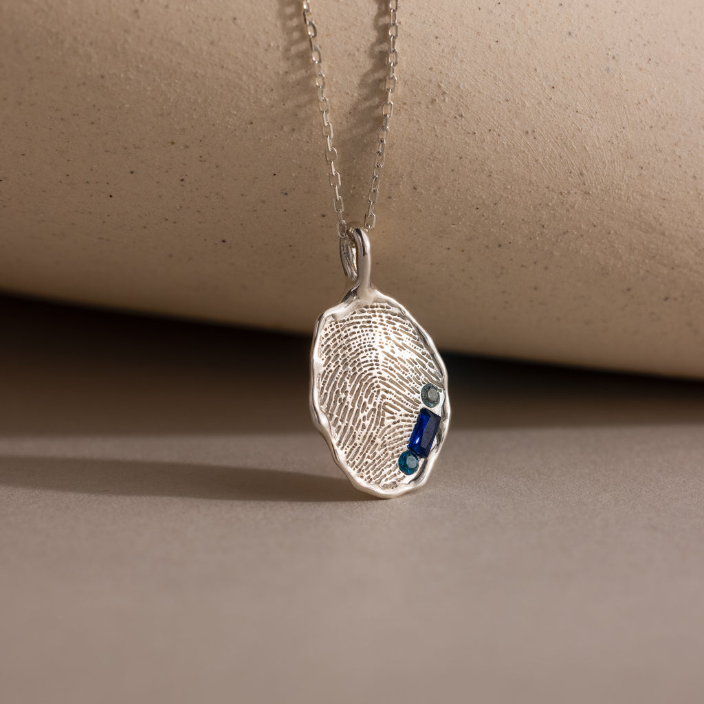 The Organic Fingerprint Multi Birthstone Necklace in Sterling Silver features an oval pendant with two small blue gemstones on a delicate chain—a personalized, elegant keepsake and perfect birthstone necklace.