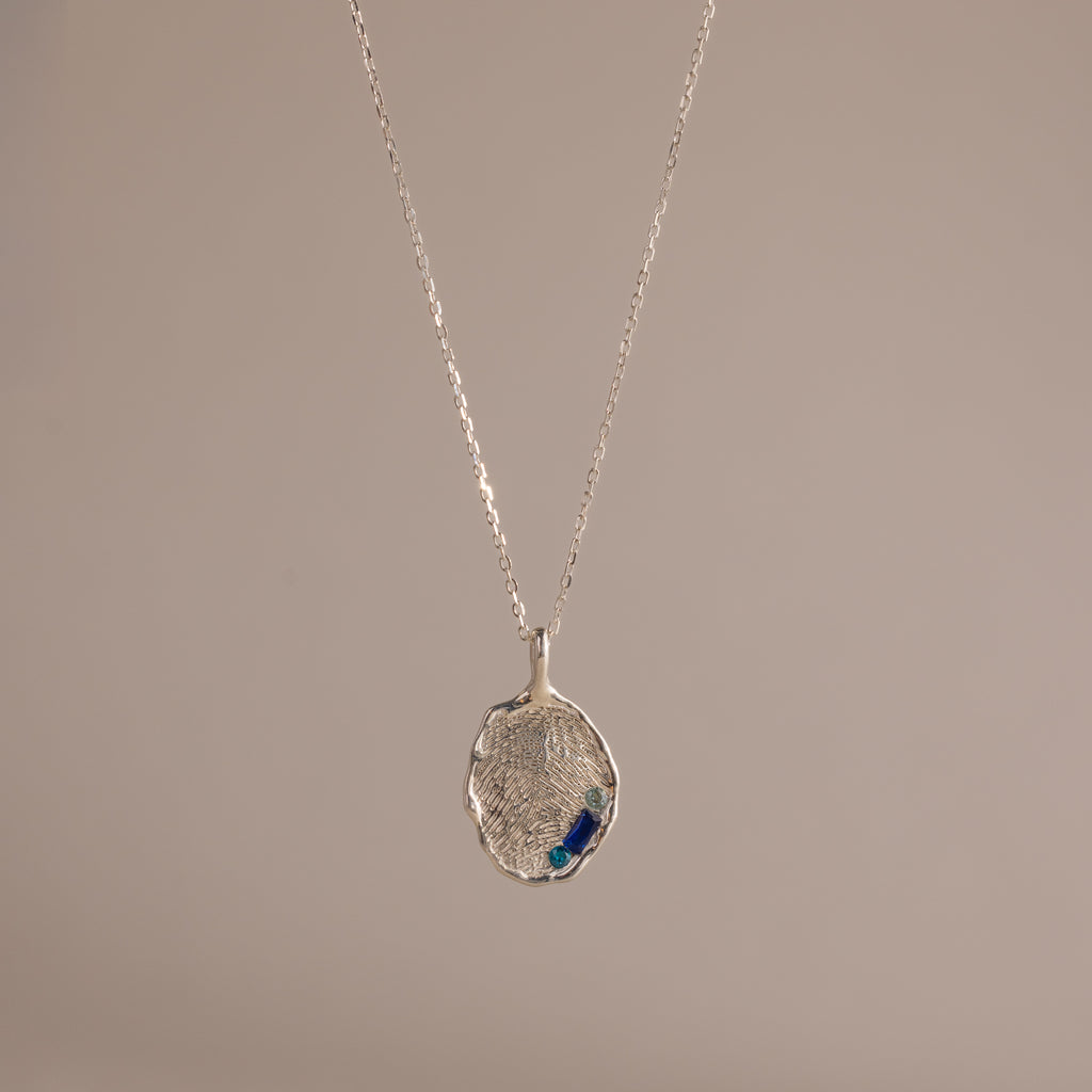 The Organic Fingerprint Multi Birthstone Necklace in Sterling Silver features a textured oval pendant with blue accents, ideal as a personalized keepsake or unique fingerprint necklace, presented on a plain beige background.