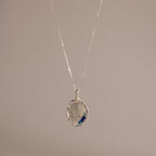 The Organic Fingerprint Multi Birthstone Necklace in Sterling Silver features a textured oval pendant with blue accents, ideal as a personalized keepsake or unique fingerprint necklace, presented on a plain beige background.