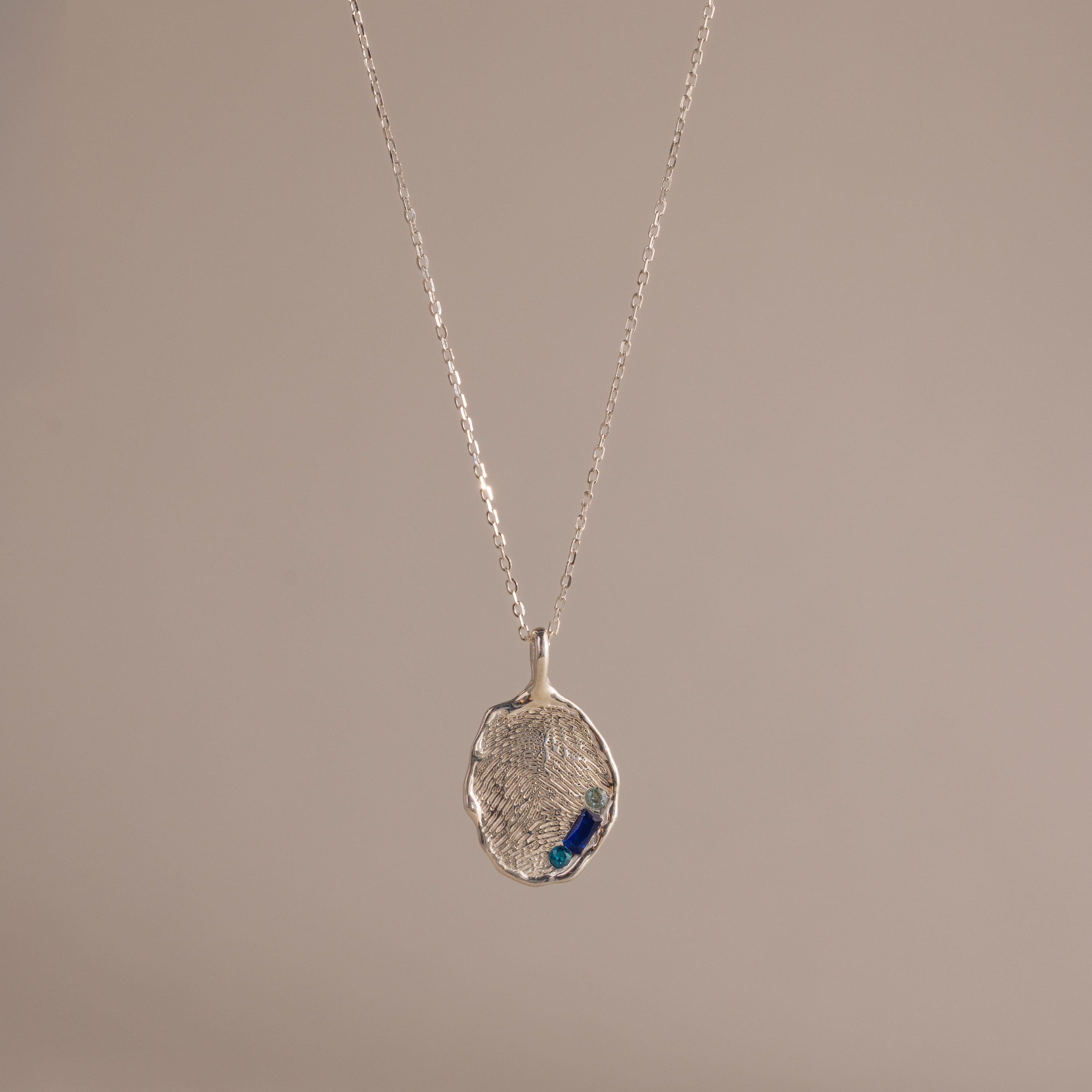 The Organic Fingerprint Multi Birthstone Necklace in Sterling Silver features a textured oval pendant with blue accents, ideal as a personalized keepsake or unique fingerprint necklace, presented on a plain beige background.