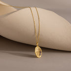 The Organic Fingerprint Multi Birthstone Necklace in 18K Gold features a textured oval charm with small gemstones, displayed on a neutral sculpted surface.