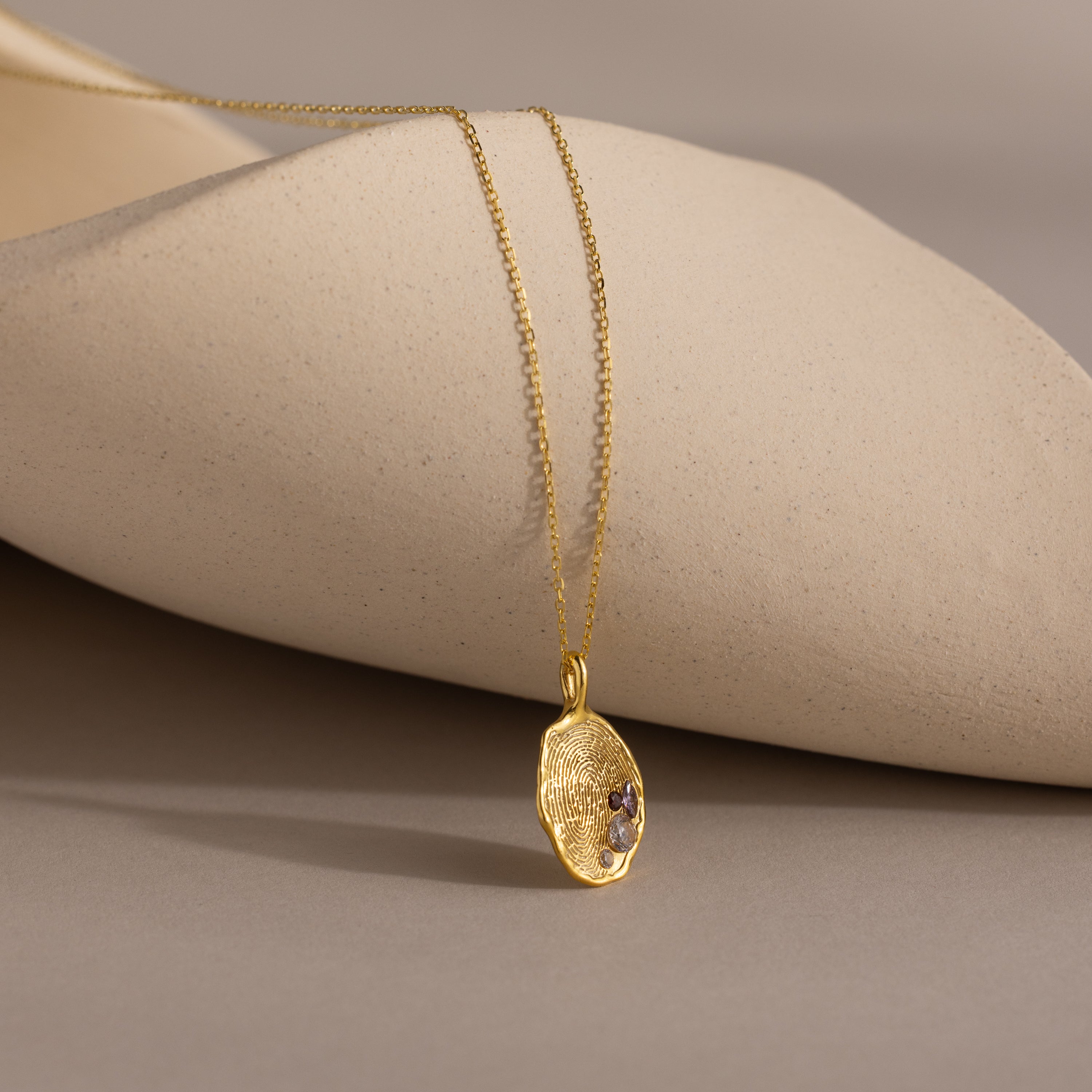 The Organic Fingerprint Multi Birthstone Necklace in 18K Gold features a textured oval charm with small gemstones, displayed on a neutral sculpted surface.