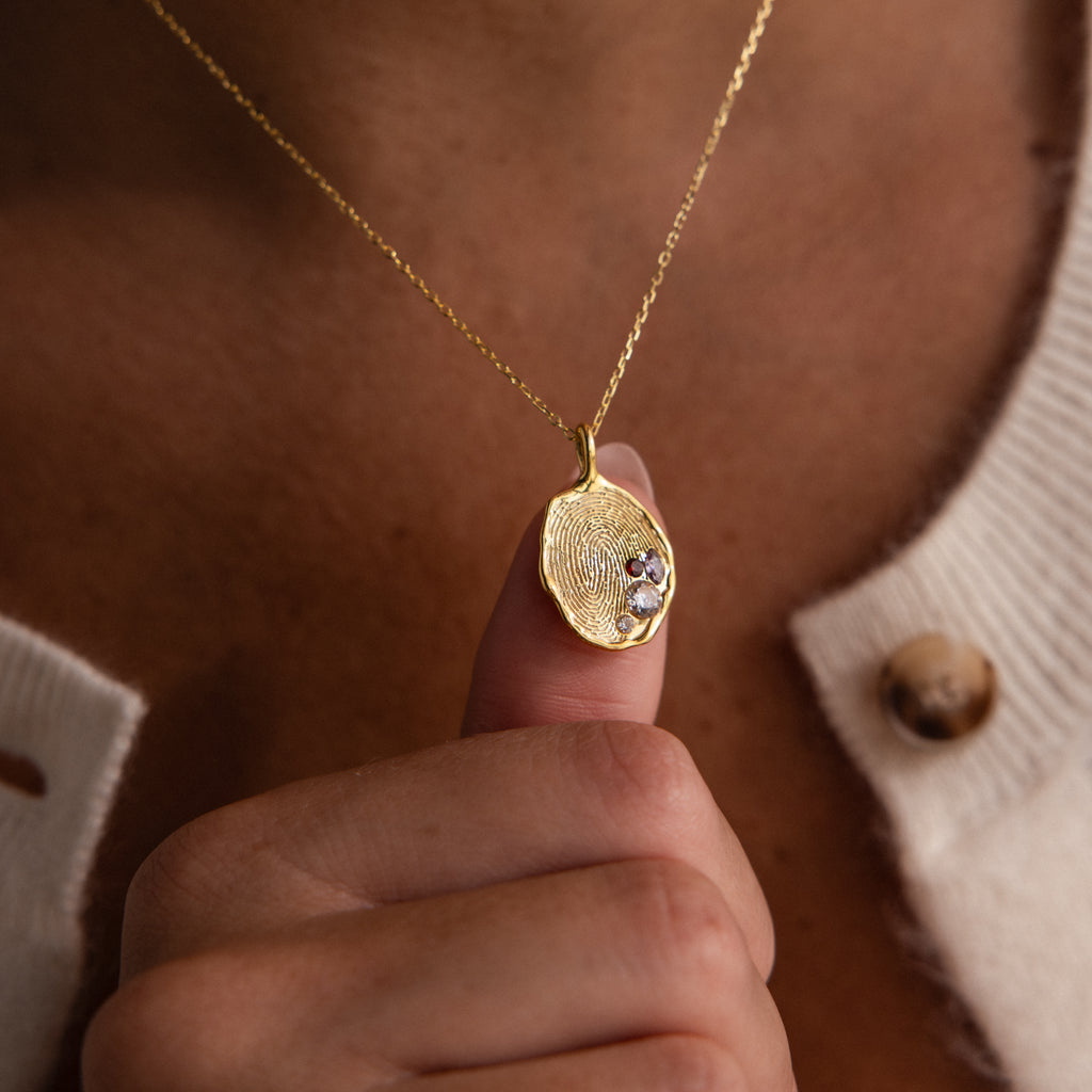 A hand holds the Organic Fingerprint Multi Birthstone Necklace—a personalized gold pendant featuring a fingerprint and two birthstones, styled over a cream sweater.