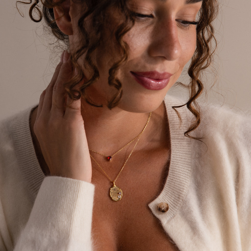 A woman in a cream cardigan touches her neck, wearing the Organic Fingerprint Multi Birthstone Necklace in Rose Gold, showcasing her love for personalized jewelry.