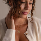 Woman with curly hair wears a cream cardigan and two gold necklaces, including the Organic Fingerprint Multi Birthstone Necklace with a red pendant.