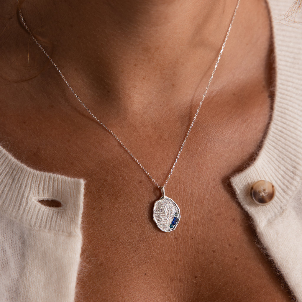 The Organic Fingerprint Multi Birthstone Necklace, featuring blue stones, is a stunning silver pendant keepsake—shown here layered over a white knit sweater for a personalized touch.
