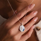 A hand with manicured nails holds an Organic Fingerprint Multi Birthstone Necklace in Sterling Silver, featuring a shell-shaped blue gemstone pendant.