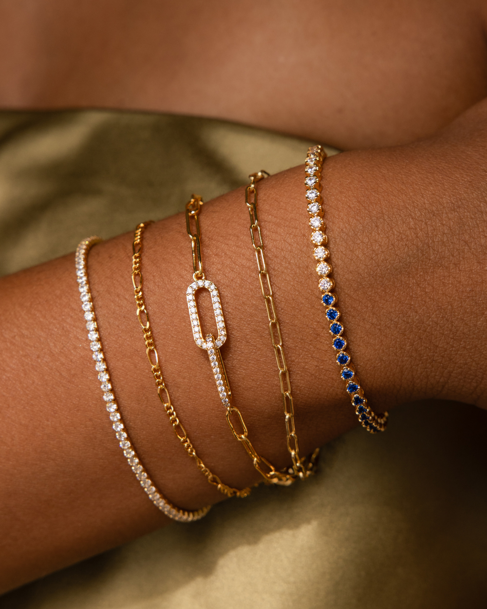 Bracelet feature photo of jewelry model wearing a gold bracelet stack on her wrist in this order: blue diamond tennis bracelet, paperclip chain bracelet, pave link pendant paperclip bracelet, figaro chain bracelet, and a diamond tennis bracelet.