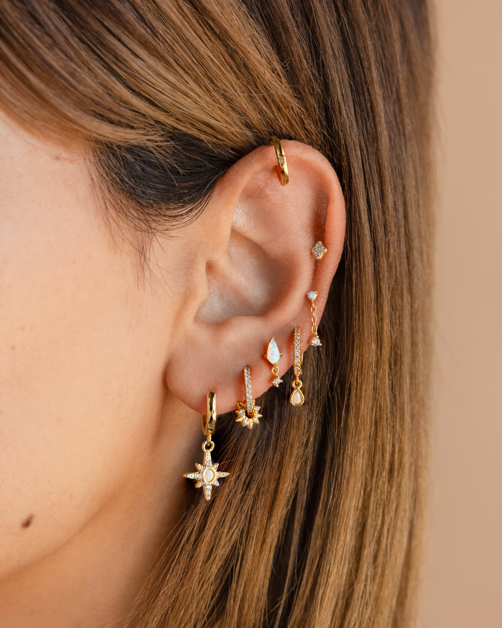 Earring feature photo of a jewelry model showcasing a gold earring stack consisting of a huggie hoop cartilage, dainty diamond stud, a pave diamond huggie hoop drop earring, 2 opal drop stud earrings, and 2 pave diamond celestial sun huggie hoop drop earrings.