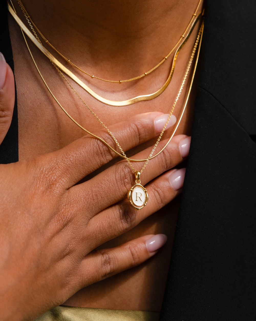 Necklace feature photo of a jewelry model showcasing a layered necklace stack consisting of a dainty gold station necklace, thin herringbone necklace, and pearl initial pendant necklace.