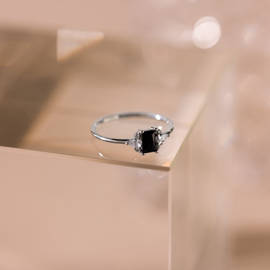 The Black Baguette Diamond Ring in Sterling Silver features a baguette-cut black diamond centerpiece with clear side stones, set on a pale, reflective surface for a vintage-inspired look.