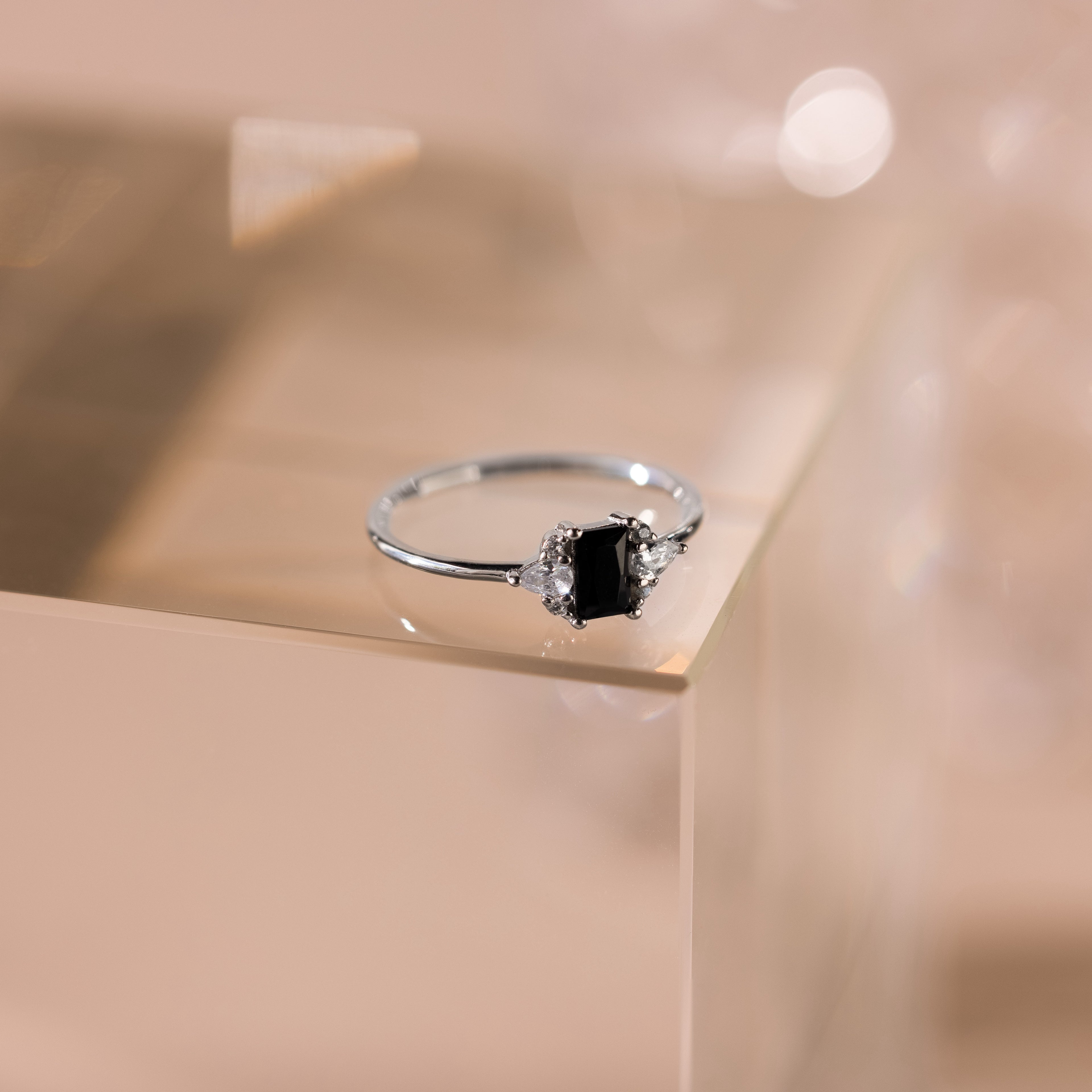 The Black Baguette Diamond Ring in Sterling Silver features a baguette-cut black diamond centerpiece with clear side stones, set on a pale, reflective surface for a vintage-inspired look.