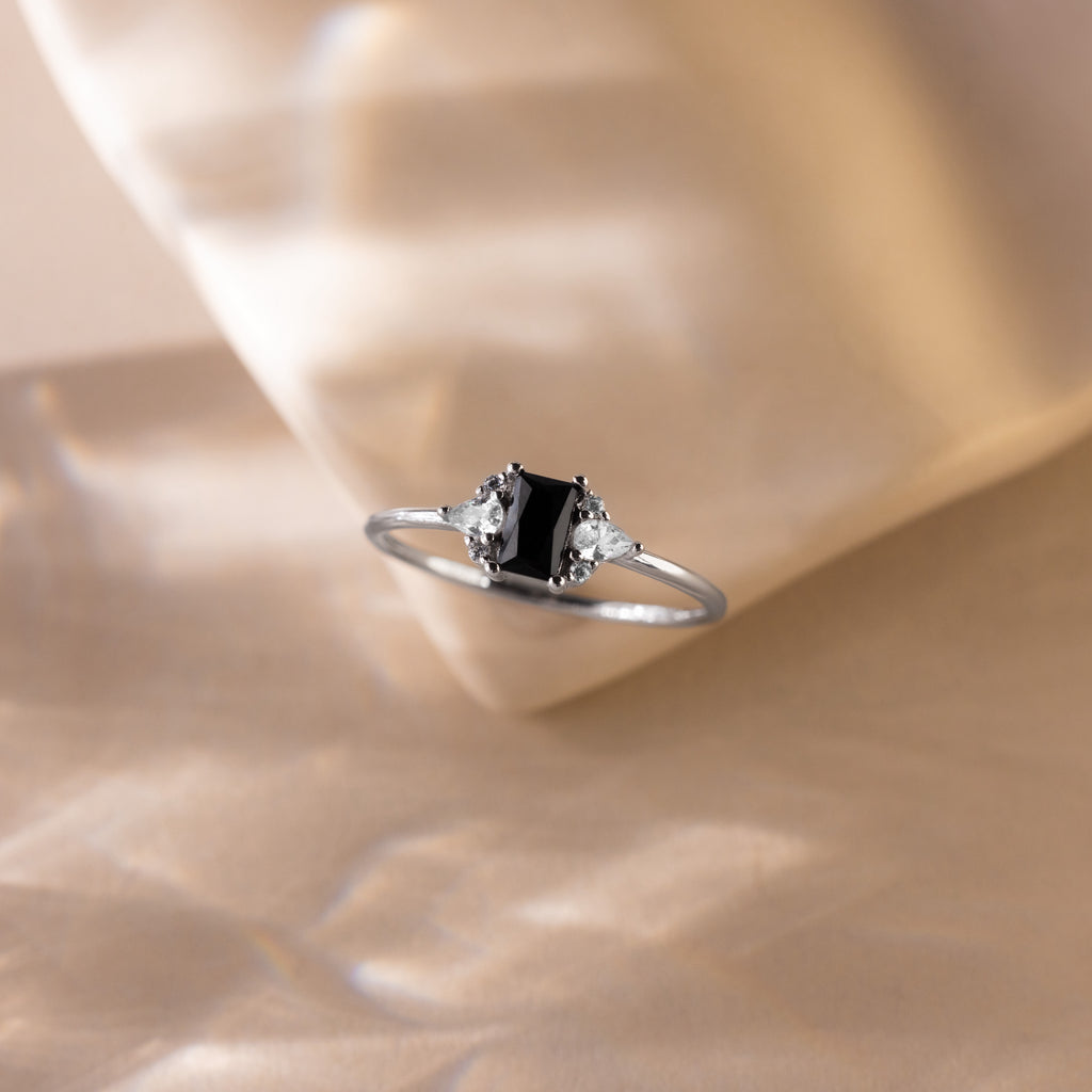 The Black Baguette Diamond Ring in Sterling Silver features a vintage-style silver band with a baguette centerpiece and two clear stones, elegantly displayed on a soft beige background.
