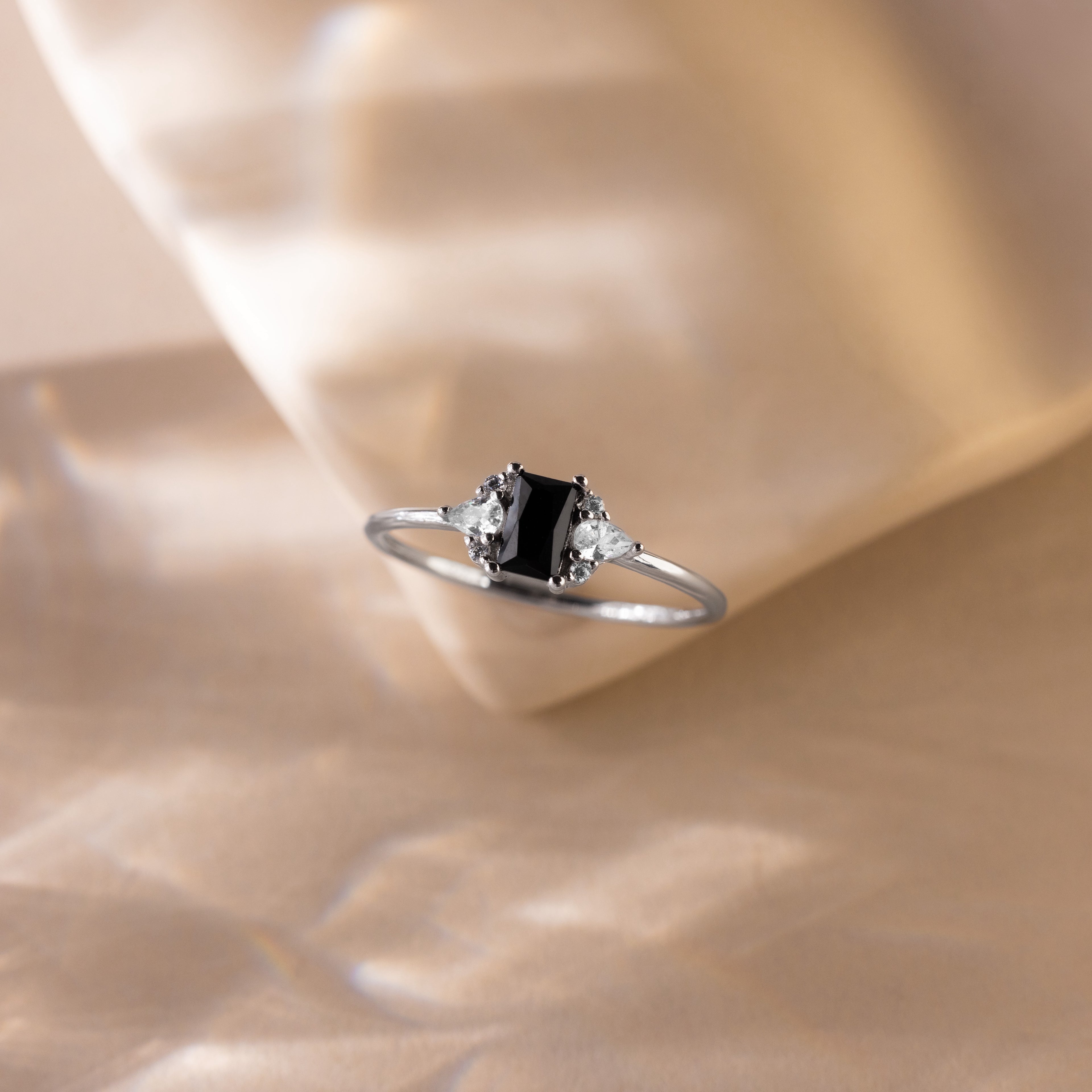 The Black Baguette Diamond Ring in Sterling Silver features a vintage-style silver band with a baguette centerpiece and two clear stones, elegantly displayed on a soft beige background.