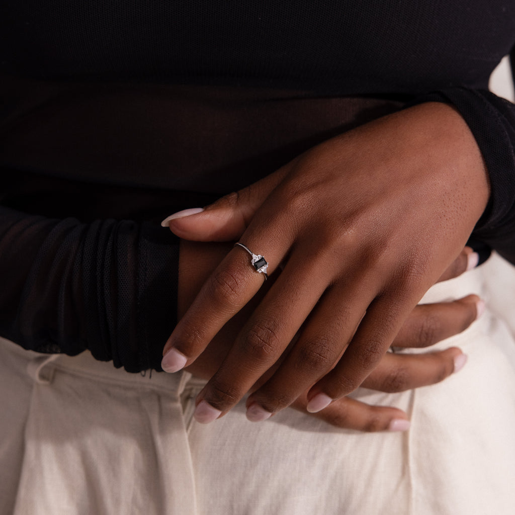A close-up of a hand with the Black Baguette Diamond Ring in Sterling Silver on the middle finger, resting on light-colored pants.