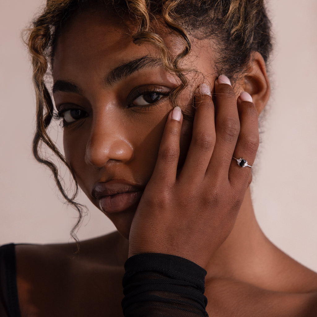 Woman with curly hair touches her face, wearing a black top and highlighting the Black Baguette Diamond Ring in Sterling Silver, featuring a striking baguette-cut centerpiece for a vintage-inspired look.